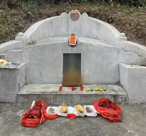 A granite grave on a cleared hillside with incense burning, small red cups, mandarin oranges, roast pork and chickens laid out in front, and red paper on top. The inscription is blurred.