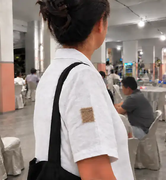 A woman at a wake in an HDB void deck in Singapore, seen from behind, with a small square of brown burlap-textured cloth pinned with a safety pin to the left sleeve of her white blouse.