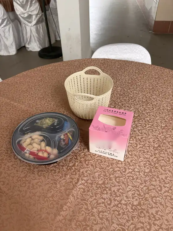 A round table at a wake with a brown patterned cloth, set with a covered tray of nuts and sweets, a small cream basket and a box of tissues.
