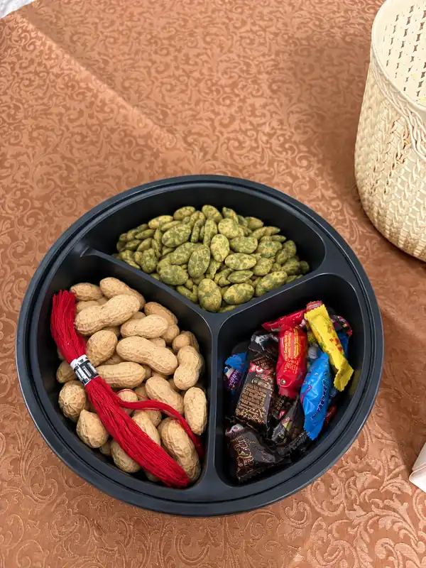 A round black tray with three compartments: peanuts in their shells with a bundle of red threads, green-coated nuts, and wrapped sweets.
